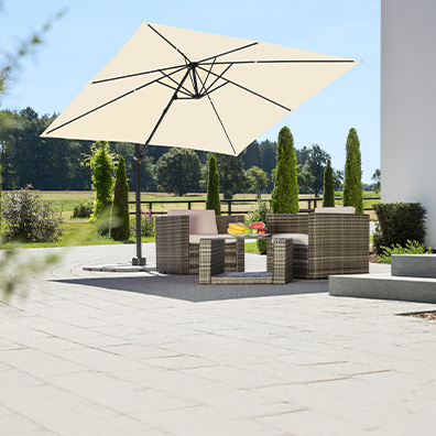 Auf einer Terrasse befinden sich Gartenmöbel und ein beiger Sonnenschirm. Im Hintergrund ist eine Wiese zusehen.